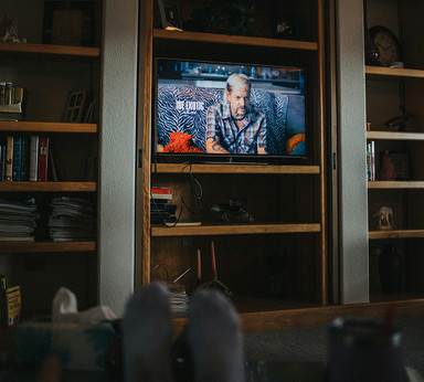 A living room with a wall of bookshelves and a television showing Joe Exotic, and the feet of someone on a couch watching the tv.