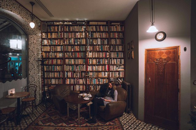 A woman reads a newspaper in a thick leather armchair in a cafe with walls covered in books.